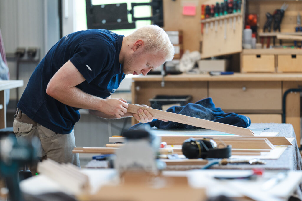 Man in a navy shirt bending over a wooden plank, sanding or shaping it in a workshop with tools and workbenches in the background